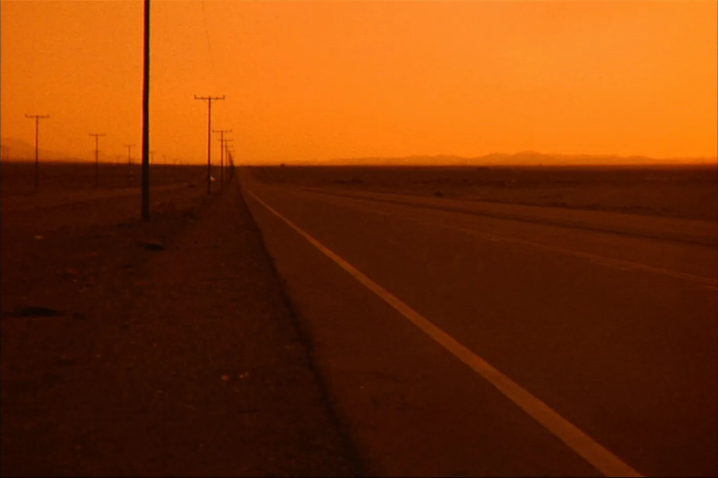 An empty two-lane road running into open desert at dusk, with distant low hills and receding telephone poles.