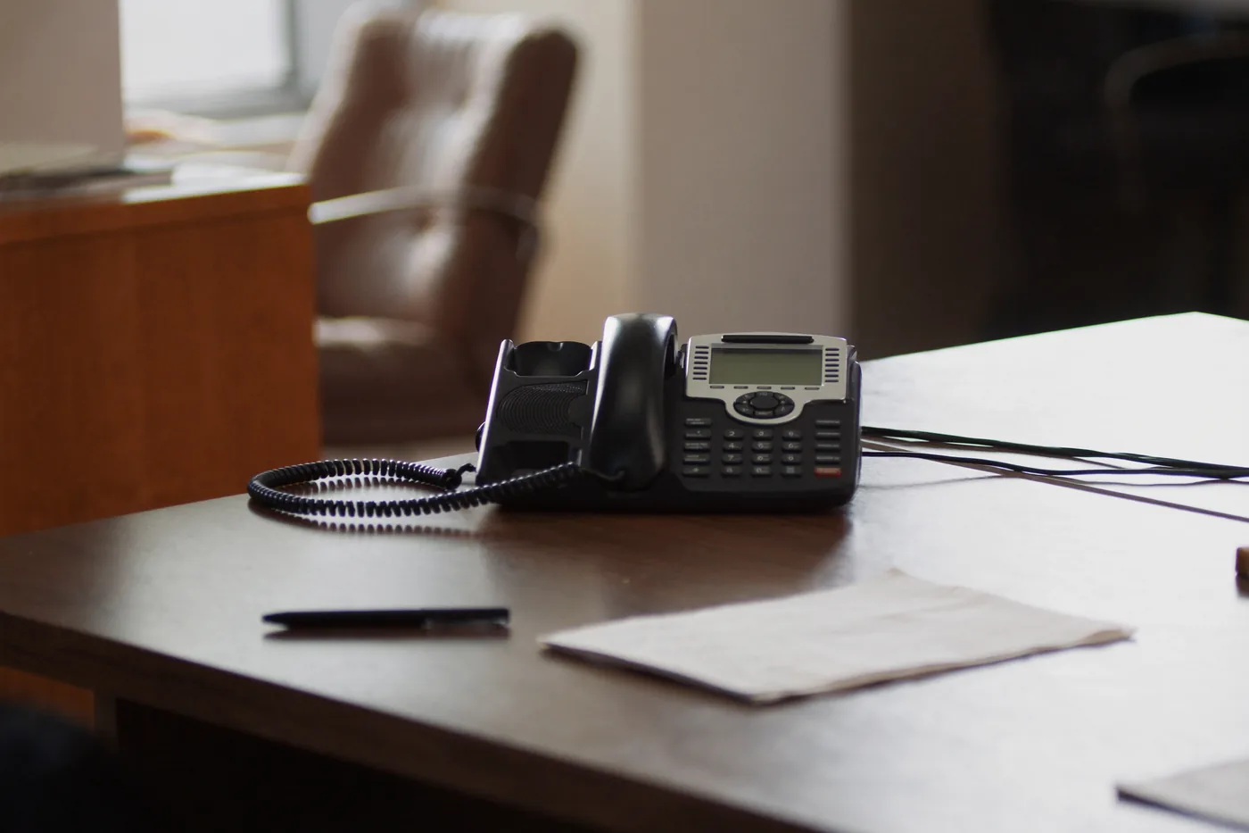 A desk telephone and a blank notepad with a pen on a wooden desk, in warm afternoon light.