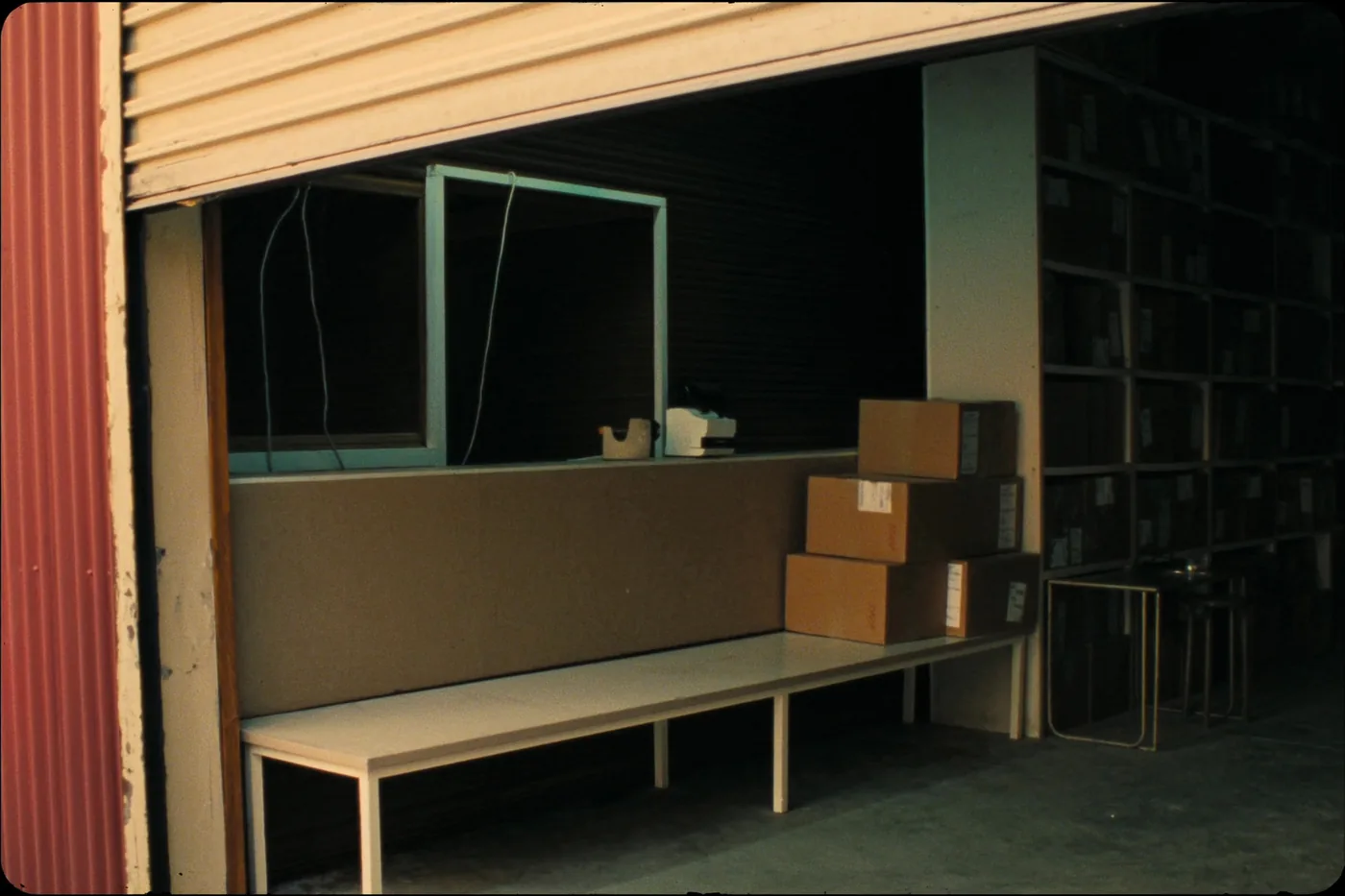 A small retailer's packing bench in a stock room: plain kraft boxes, a tape dispenser, an unbranded label printer and metal shelving.