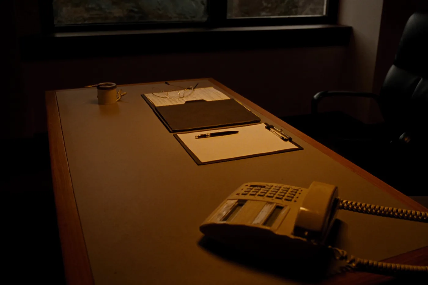 A small professional office desk in warm window light: a closed folder, reading glasses, a blank notepad and a desk phone.