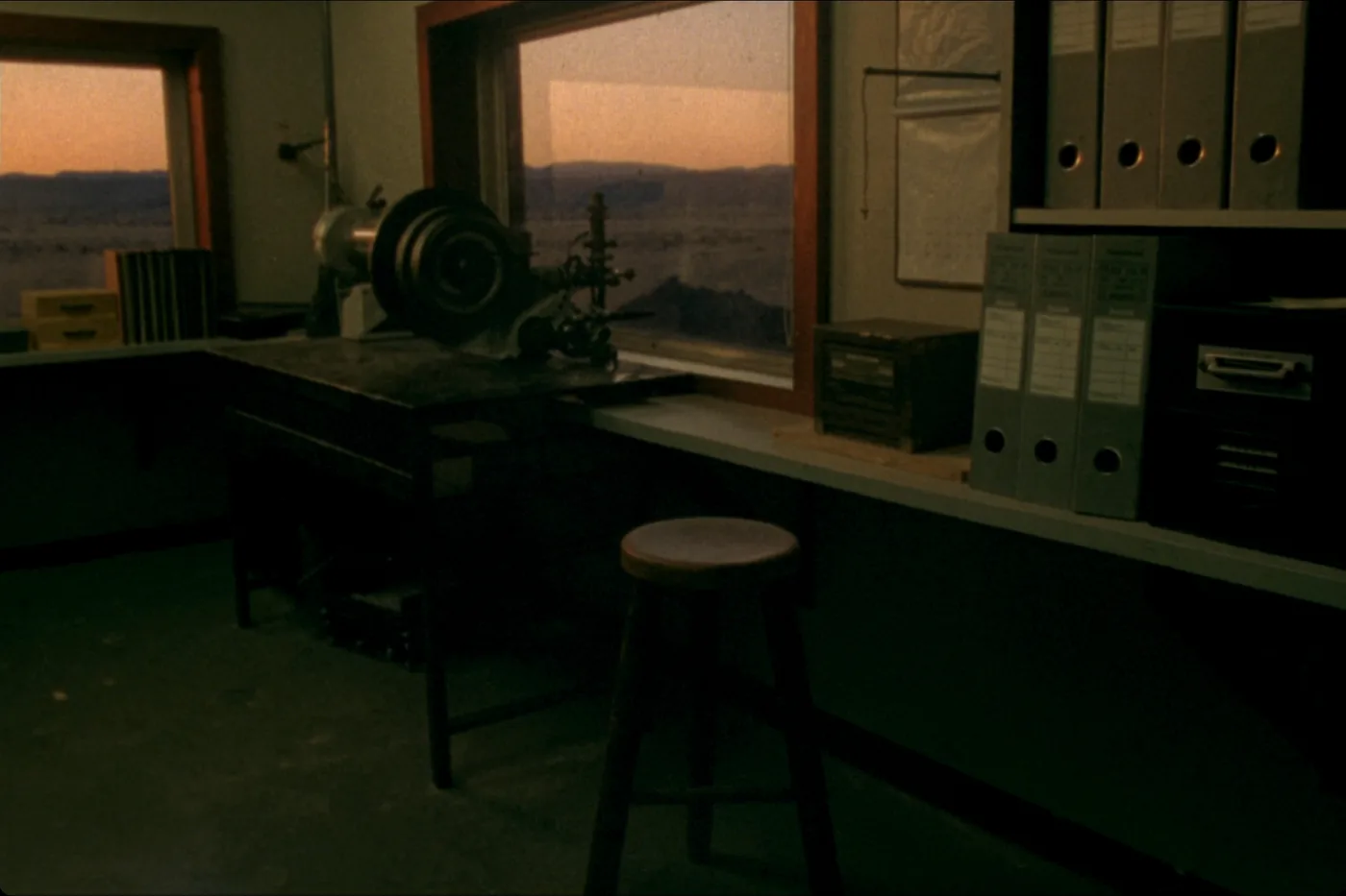 A quiet corner of a small workshop office at dusk: a shelf of plain boxes, a wooden stool and a window full of warm sky.